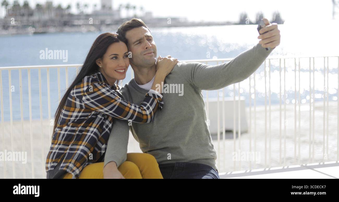 Un couple sur leur téléphone mobile selfies à la station assise sur une promenade au bord de l'smiling for the camera Banque D'Images