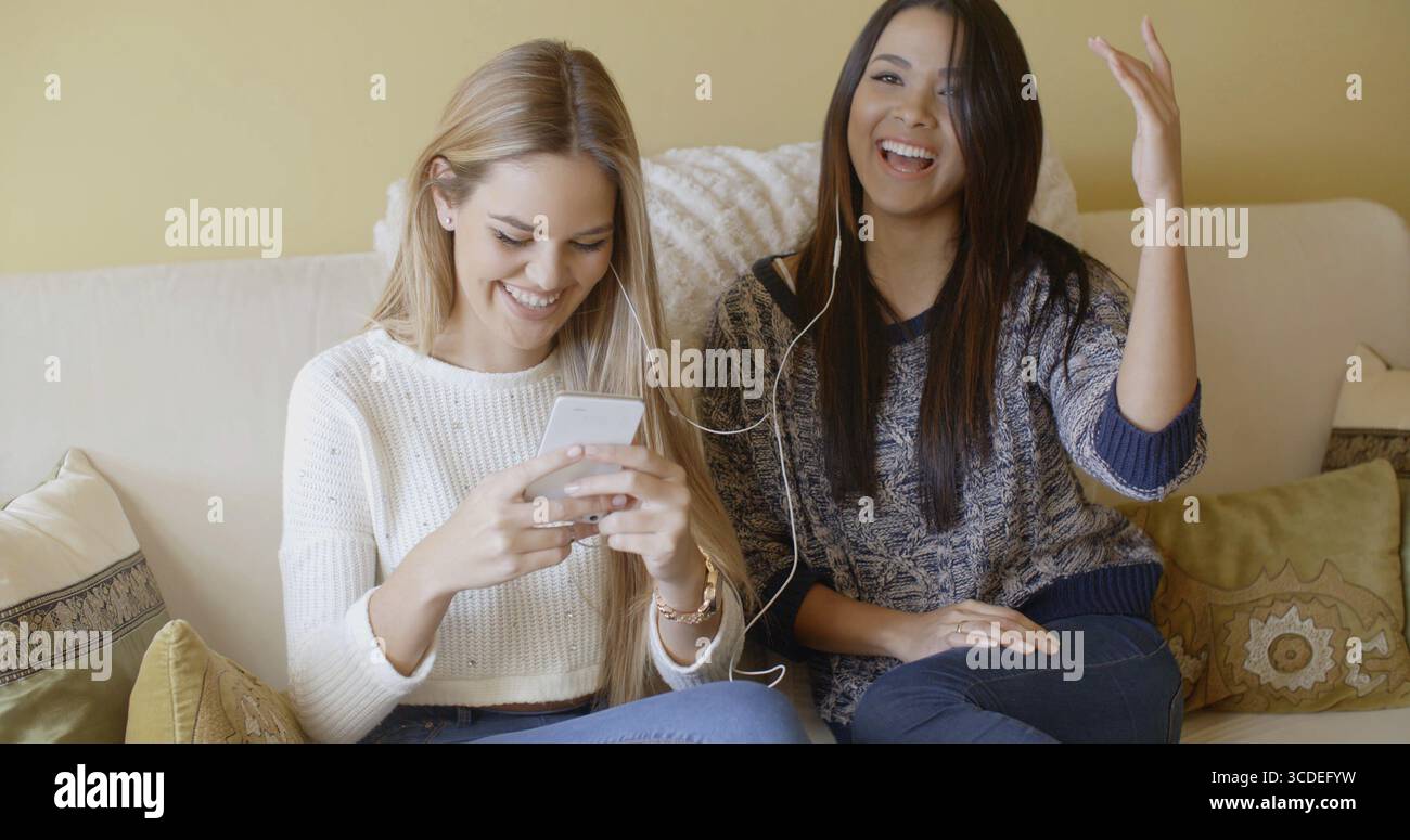 De jeunes femmes heureuses assises sur un canapé à la maison, encourageant les bonnes nouvelles qu'elles viennent de recevoir via un message texte sur un téléphone portable Banque D'Images