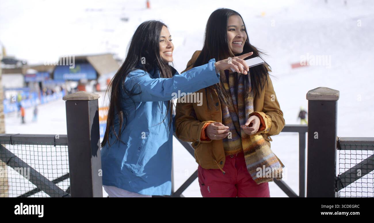 Deux jeunes femmes en vacances de ski d'hiver debout sur un balcon d'un chalet à la station souriant en pointant sur le côté Banque D'Images