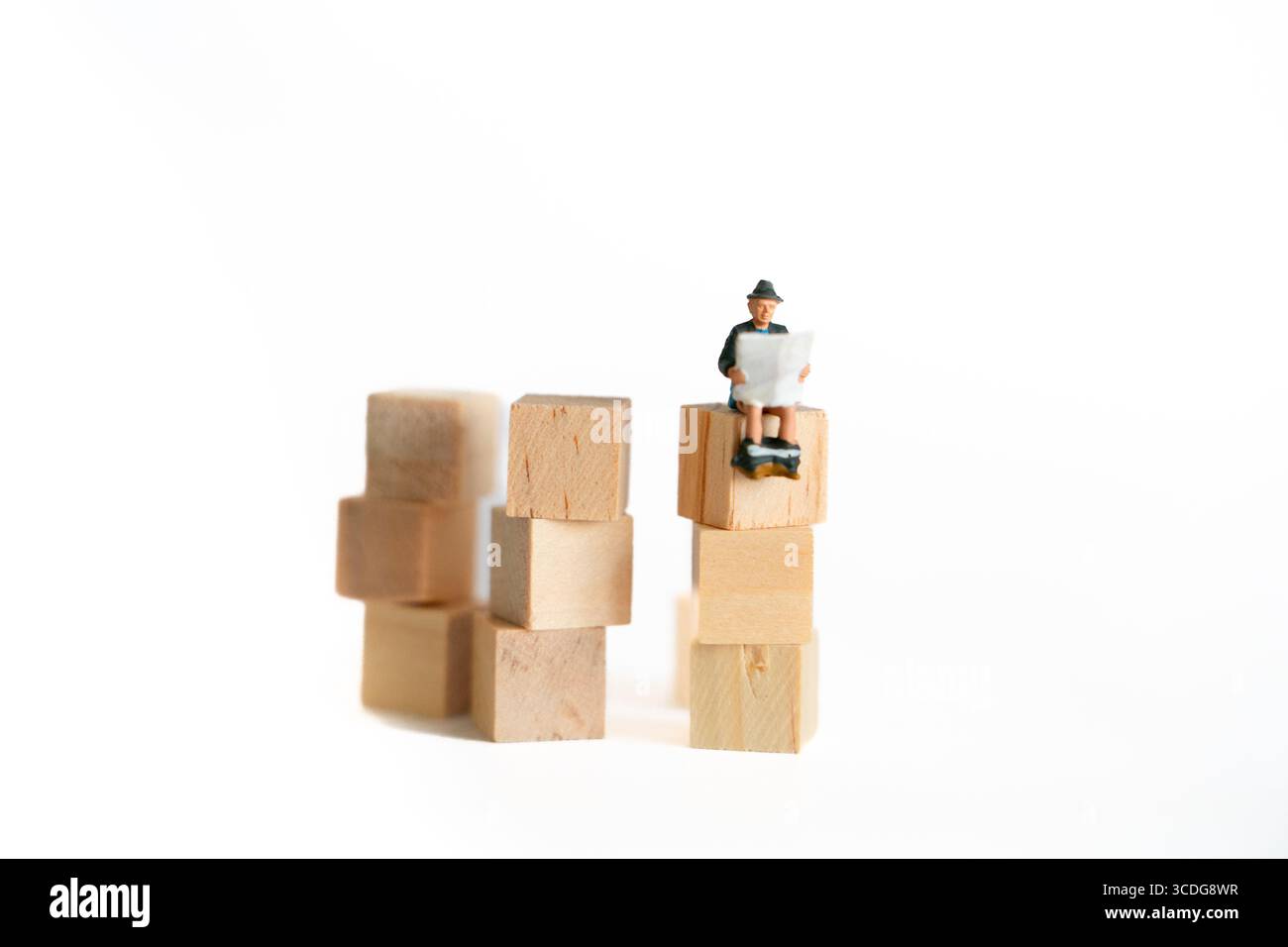 Drôle homme miniature assis sur des blocs de bois lisant le journal – scène conceptuelle fantaisiste de l'humour de toilette et de l'imagination dans l'art. Banque D'Images