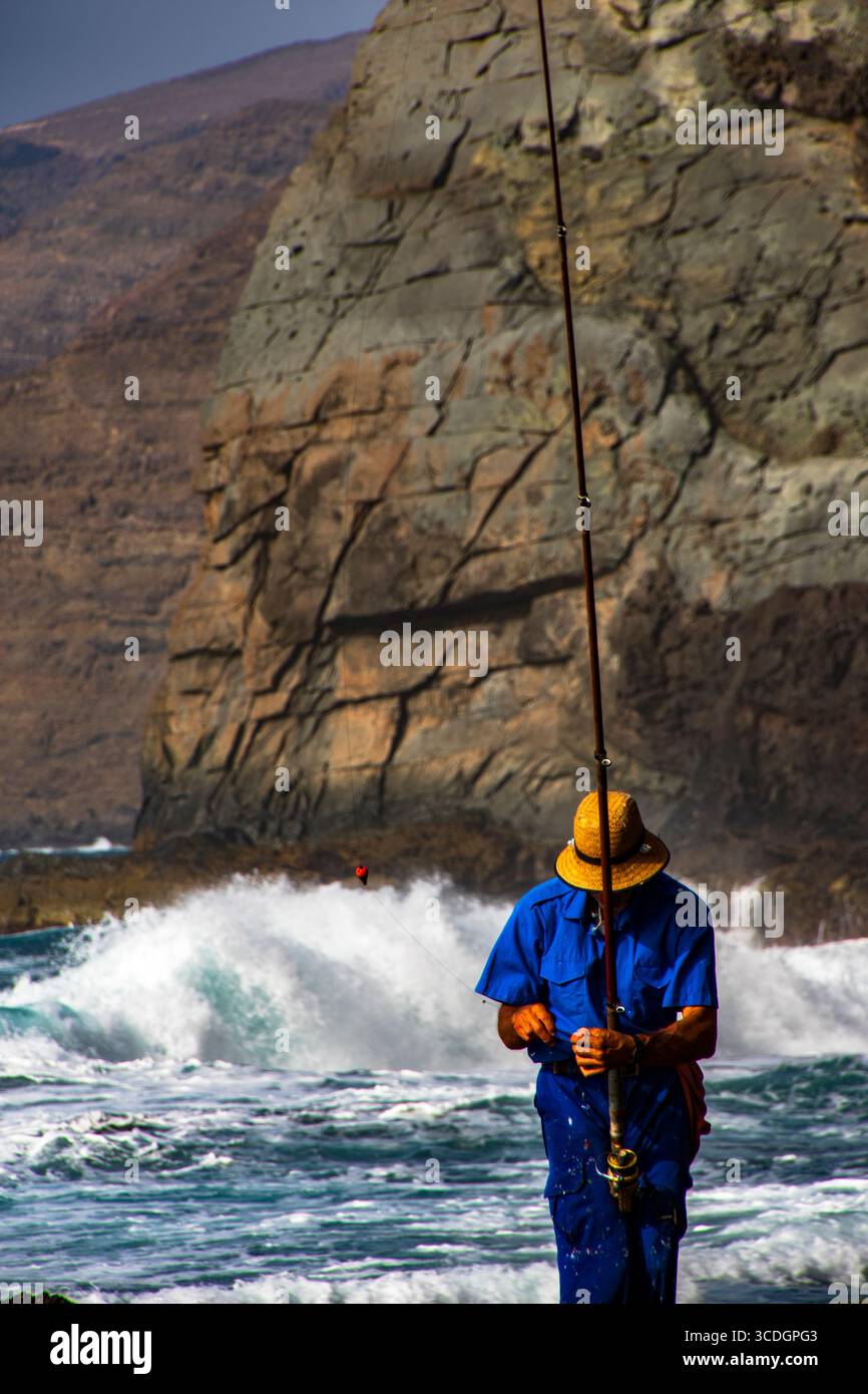 Hombre pescador con sombrero de paja y ropa azul, sobre un acantilado volcánico oscuro, practicando pesca tradicional en solitario. Banque D'Images