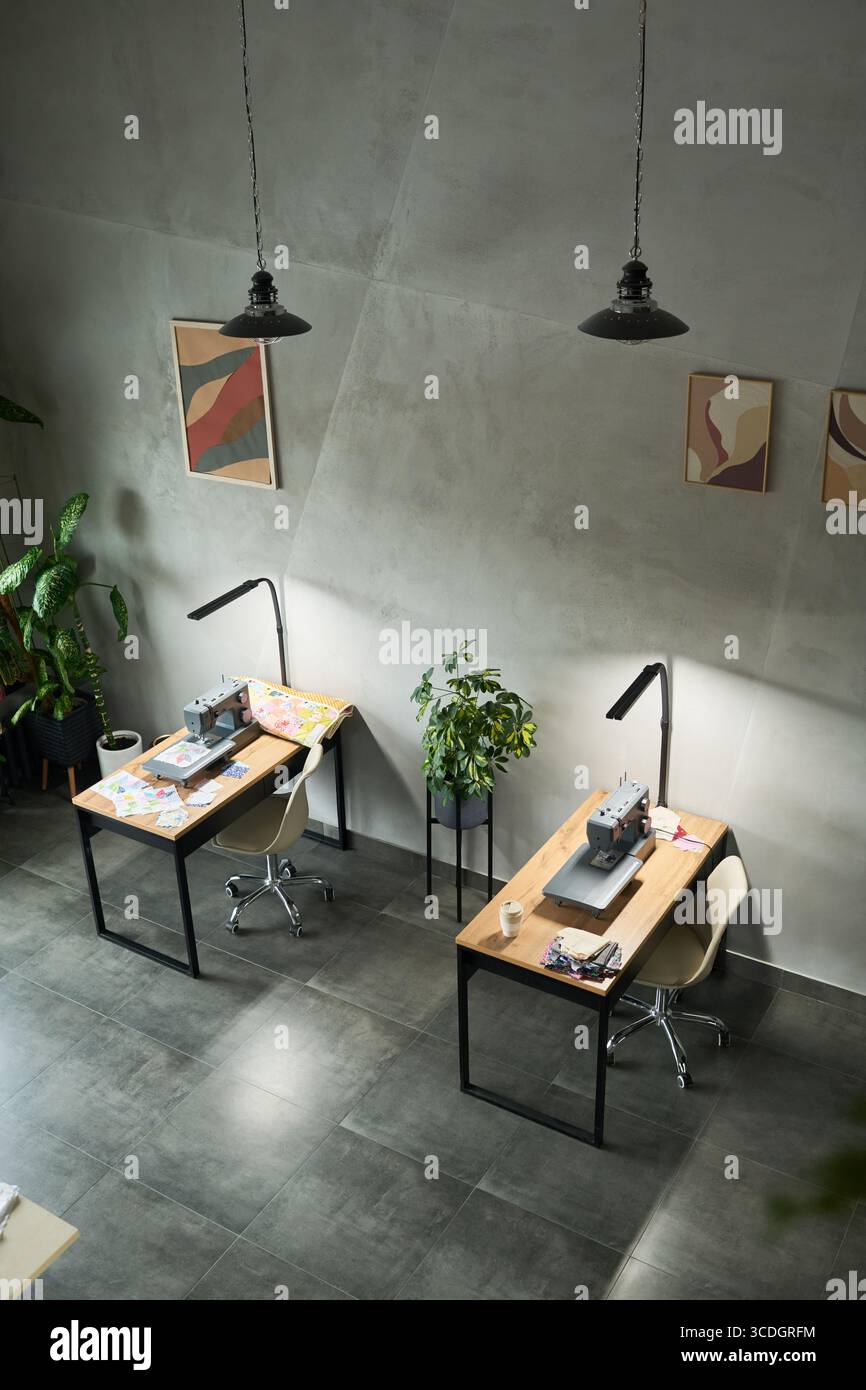 Espace de travail moderne vide montrant deux machines à coudre sur des bureaux en bois avec des morceaux de tissu, chaises ergonomiques, plantes en pot, art mural abstrait, suspensions plafonnières, carrelage Banque D'Images