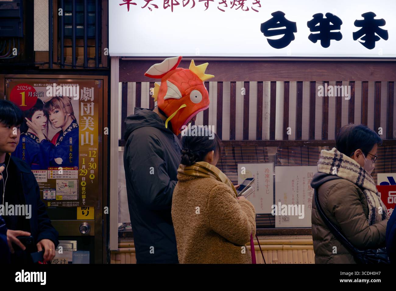 Homme portant un chapeau Magikarp ludique tout en faisant la queue à Asakusa, aux côtés d'autres personnes vérifiant leurs téléphones Banque D'Images