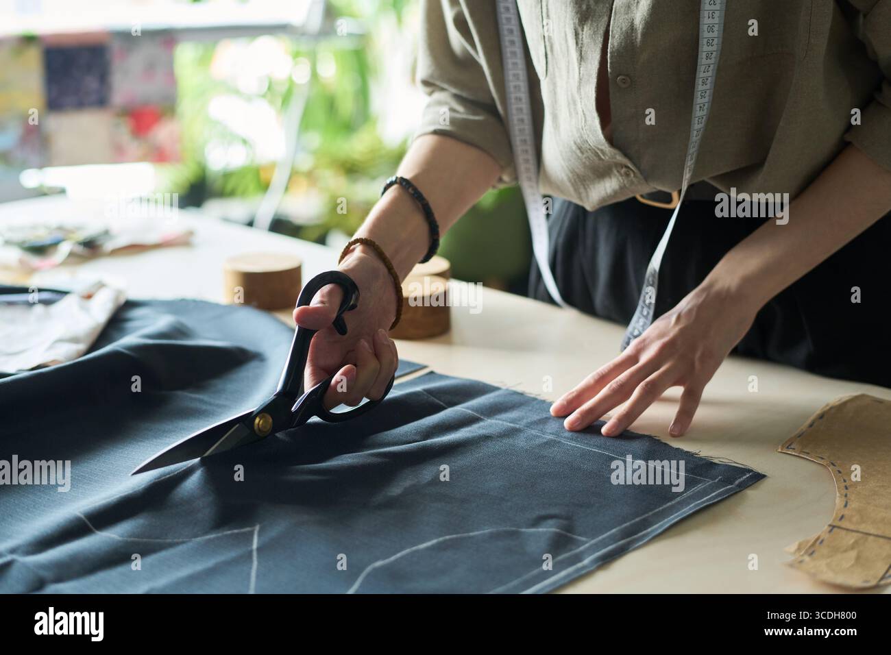 Femme jeune adulte caucasienne coupant le tissu avec de grands ciseaux sur la table de travail, ruban à mesurer autour du cou, mains guidant soigneusement le textile, engagé dans le processus de fabrication de vêtements Banque D'Images