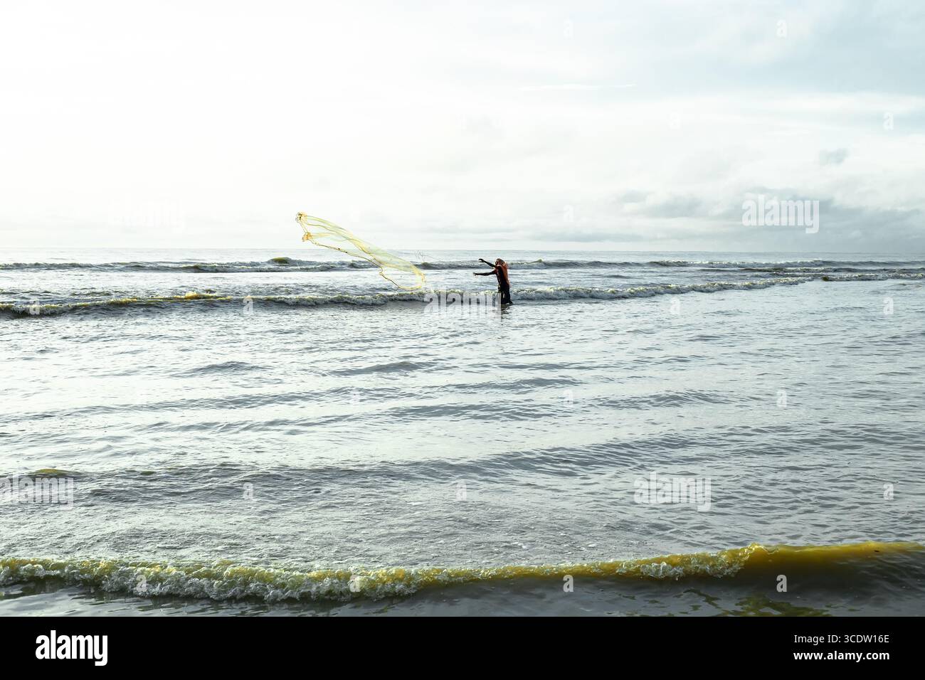 Cox's Bazar, Bangladesh - 11 octobre 2019 : vue d'un pêcheur solitaire pataugant dans les vagues, jetant un filet jaune éclatant contre les tons doux de la mer et du ciel. Banque D'Images