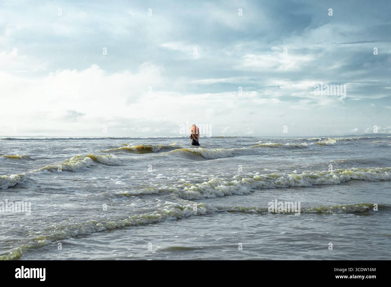 Cox's Bazar, Bangladesh - 11 octobre 2019 : vue d'un personnage solitaire dans une veste orange se tient au milieu des vagues turbulentes de la baie du Bengale, sous un vaste ciel parsemé de nuages. Banque D'Images
