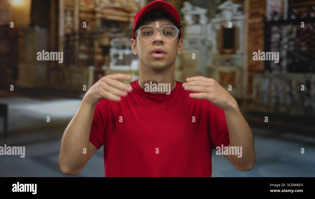 Jeune homme hispanique portant un chapeau rouge et un t-shirt rouge fait un geste de signe OK avec les mains dans le bâtiment ; approbation calme. Banque D'Images