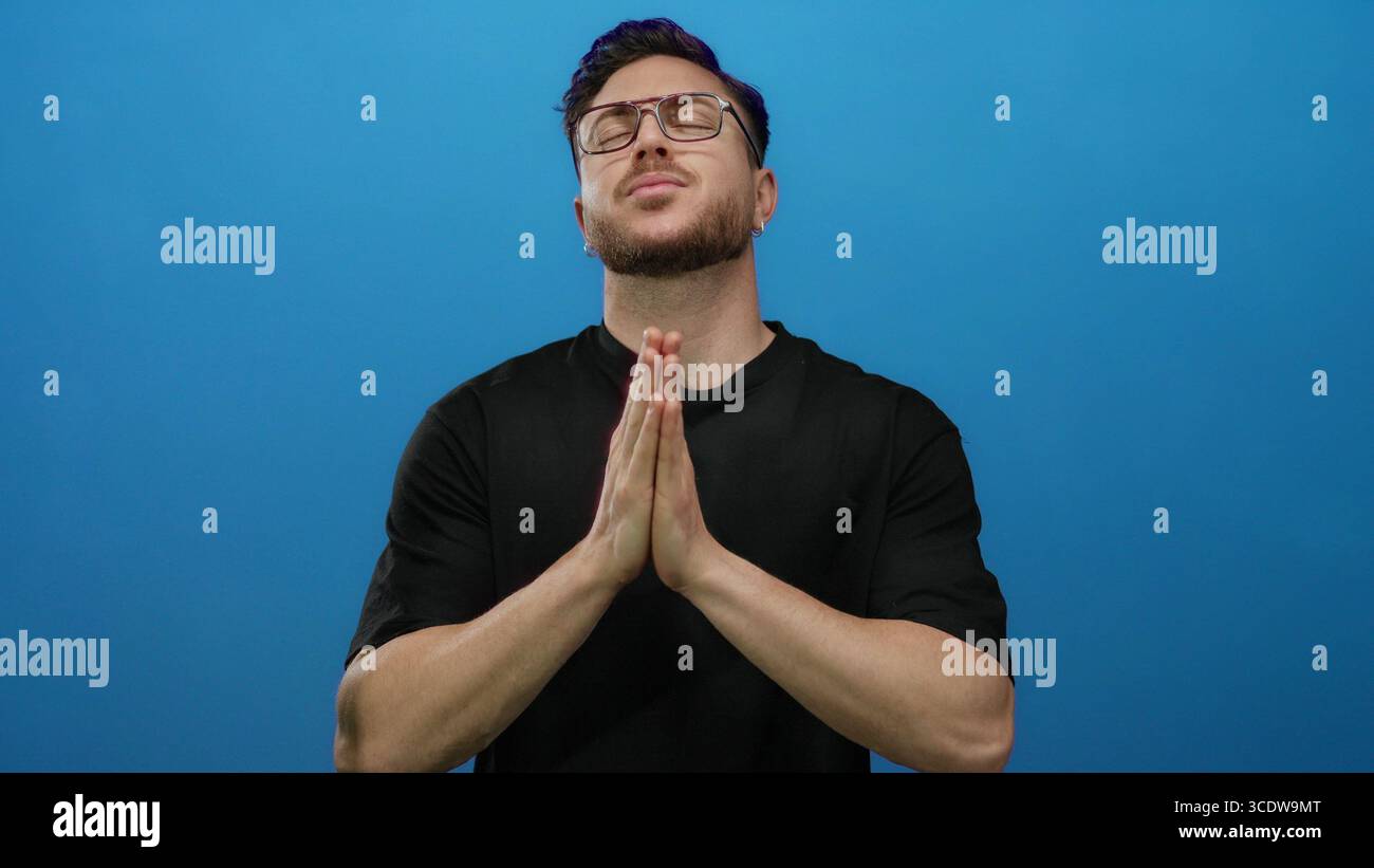 Jeune homme à la barbe priant les yeux fermés sur fond bleu, exprimant espoir et spiritualité dans un cadre isolé. Banque D'Images