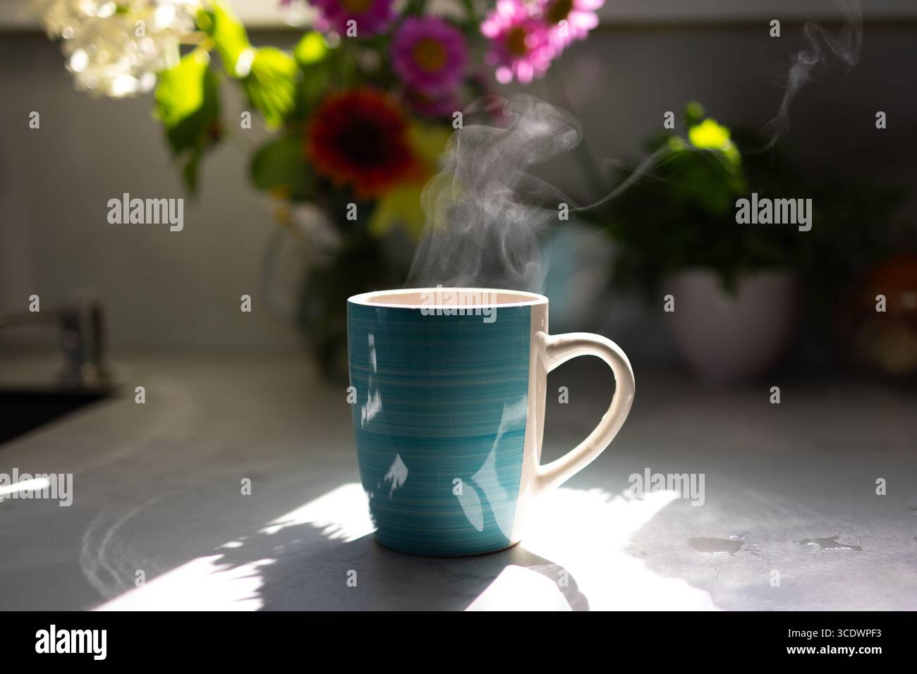 Une tasse de café fumant se trouve sur un comptoir de cuisine, éclairé par la lumière du soleil. Des fleurs colorées décorent le fond, créant une atmosphère chaleureuse et accueillante remplie d'énergie matinale. Banque D'Images