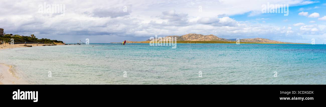 Large vue panoramique sur la plage de la Pelosa en Sardaigne, avec des eaux turquoises peu profondes, un rivage sablonneux et des collines rocheuses sous un ciel partiellement nuageux. Banque D'Images