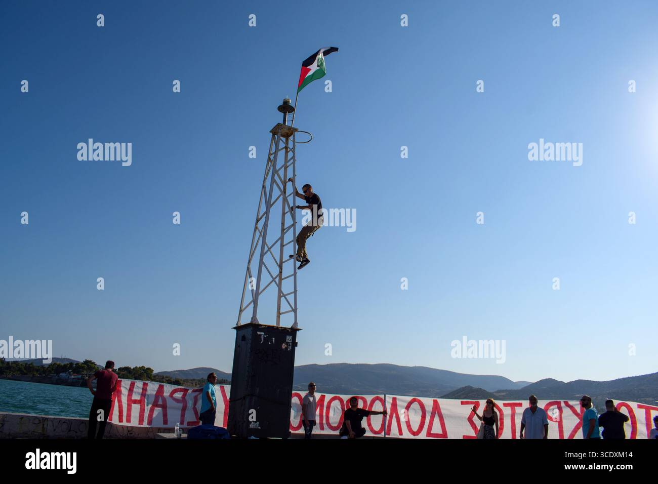 Volos, Grèce, 13 août 2025. Un manifestant accroche un drapeau palestinien sur le lampadaire du port tandis que les manifestants tiennent des banderoles avec des slogans lors d'une manifestation contre l'arrivée du navire de croisière israélien Crown Iris. Des manifestations ont eu lieu sur les îles grecques et les ports continentaux le long de la route de l'Iris de la Couronne, qui transporterait des soldats israéliens en vacances en Grèce. Crédit : Dimitris Aspiotis/Alamy Live News Banque D'Images