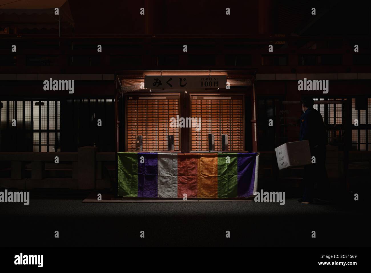 Ouvrier portant une boîte à côté du stand de fortune omikuji illuminé au temple Sensō-ji à Asakusa Banque D'Images