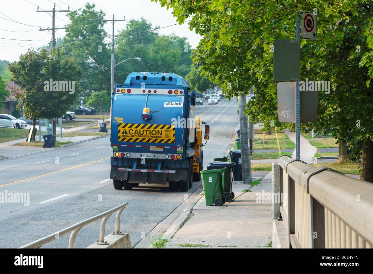 Un camion à ordures s’arrête près de bacs verts dans une rue résidentielle bordée d’arbres et de poteaux électriques. Banque D'Images