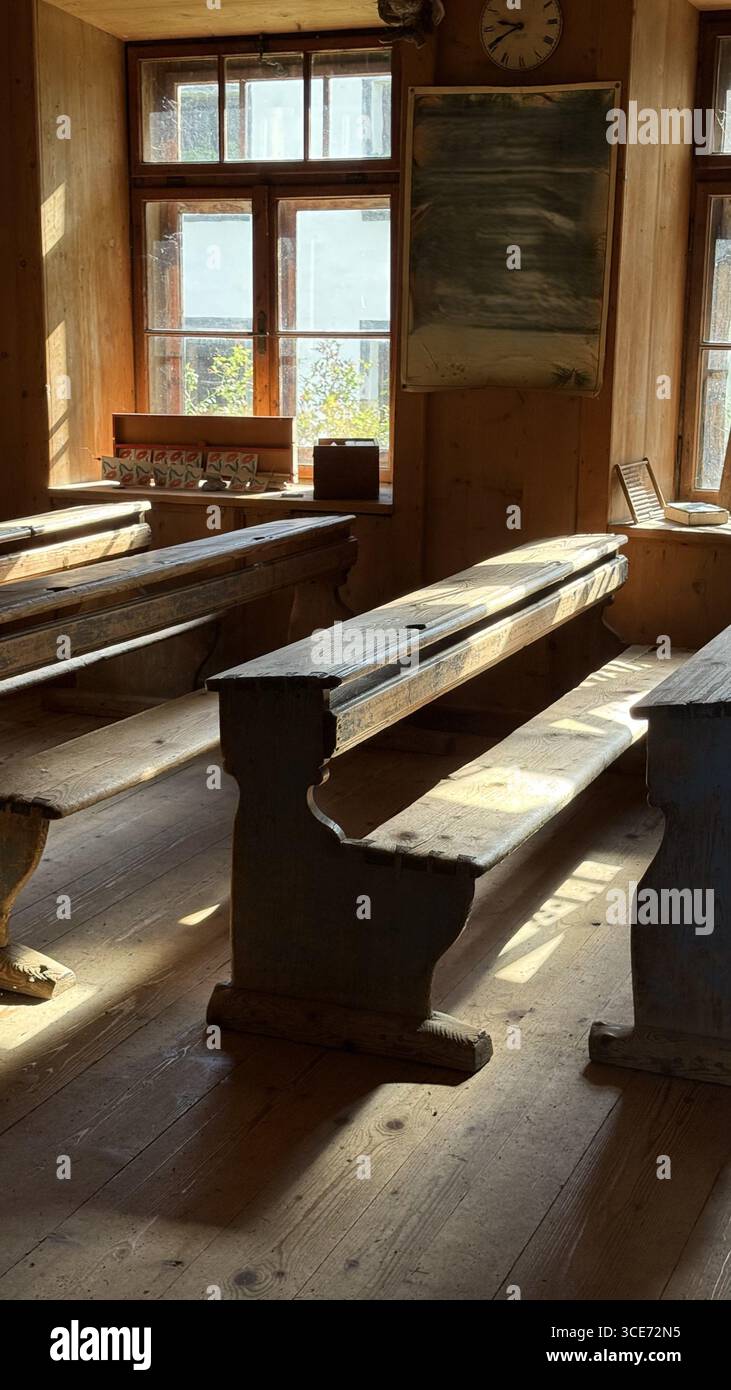 De vieux bureaux et bancs en bois dans une salle de classe d'école de village rustique évoquent un sentiment de nostalgie. La lumière du soleil filtre à travers les grandes fenêtres, mettant en évidence le Banque D'Images