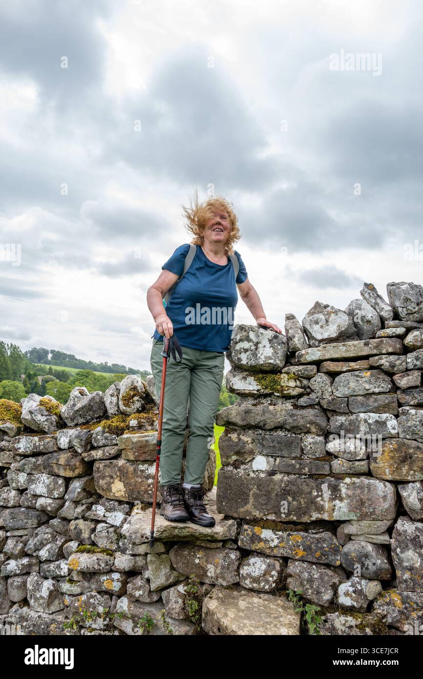 Femme grimpant sur un stile de pierre construit dans un mur de pierre sèche construit à partir de calcaire local le long de Pennine Way, Yorkshire Dales. Banque D'Images
