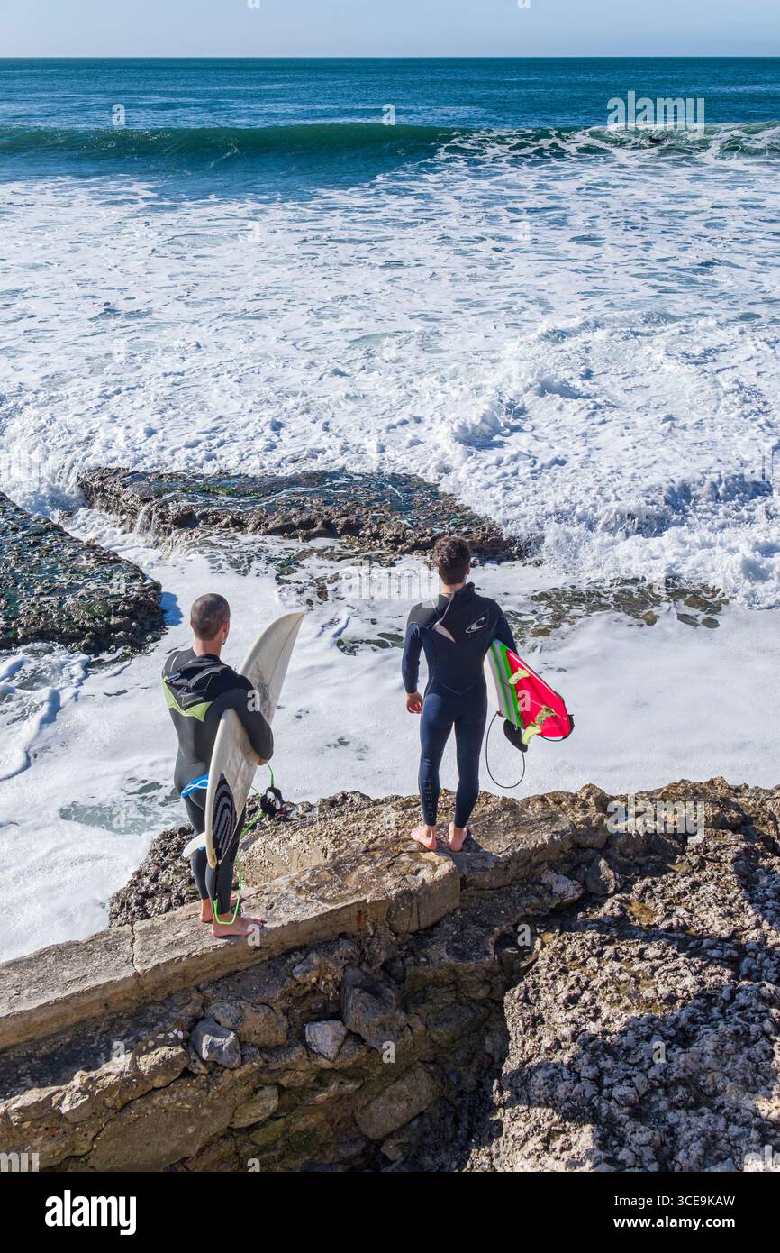 Deux jeunes hommes portant des surfeurs combinaison néoprène holding surfboards à la recherche d'un chemin d'accès à l'océan, Estoril, Lisbonne, Portugal Banque D'Images
