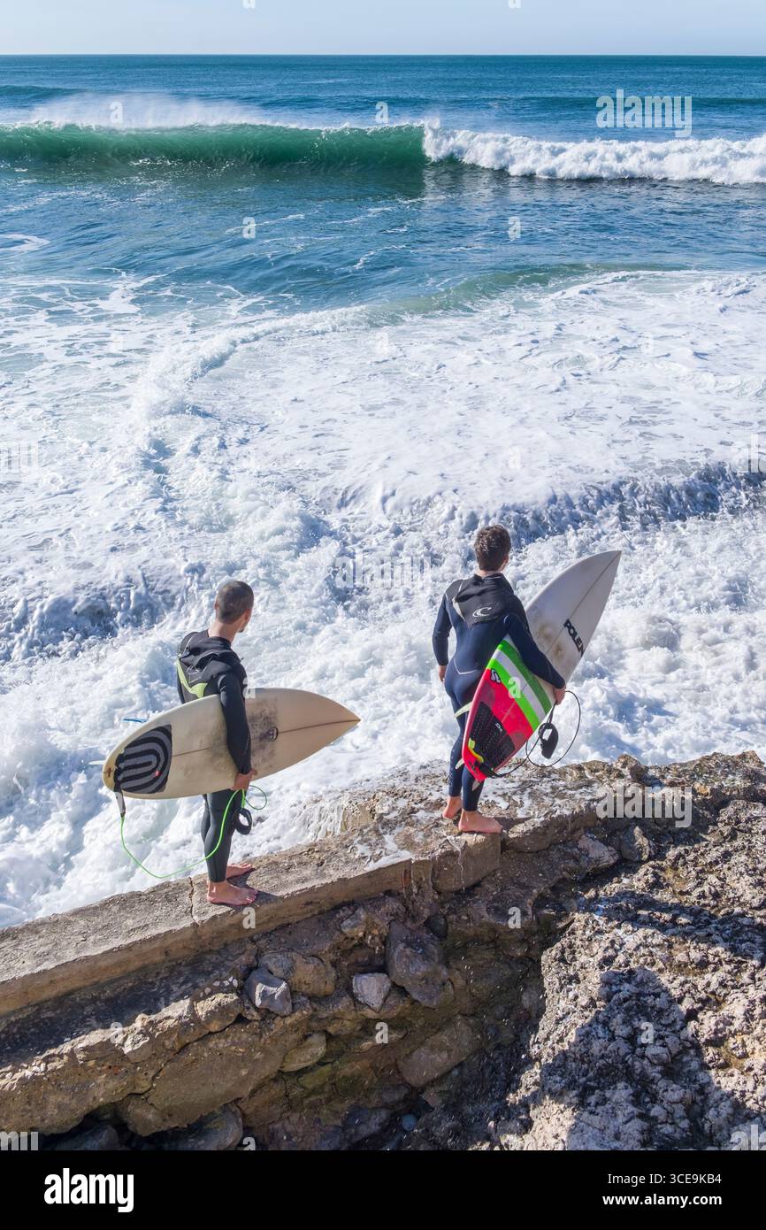 Deux jeunes hommes portant des surfeurs combinaison néoprène holding surfboards à la recherche d'un chemin d'accès à l'océan, Estoril, Lisbonne, Portugal Banque D'Images