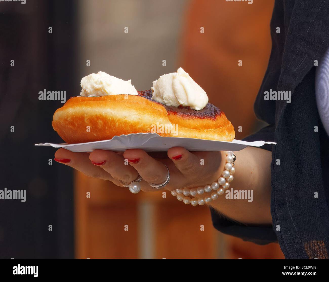 Womans main tient un plateau de papier avec deux pâtisseries frites surmonté de crème fouettée, bracelet de perles et ongles rouges visibles, photographié à un fo extérieur Banque D'Images