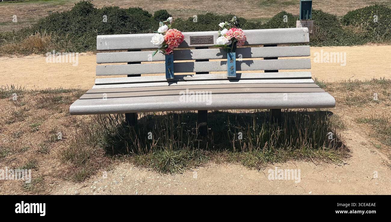 Un banc commémoratif avec des arrangements floraux placés dans des vases attachés, debout le long d'un sentier de randonnée dans un parc par une journée ensoleillée brillante - Image de stock capturée avec un smartphone