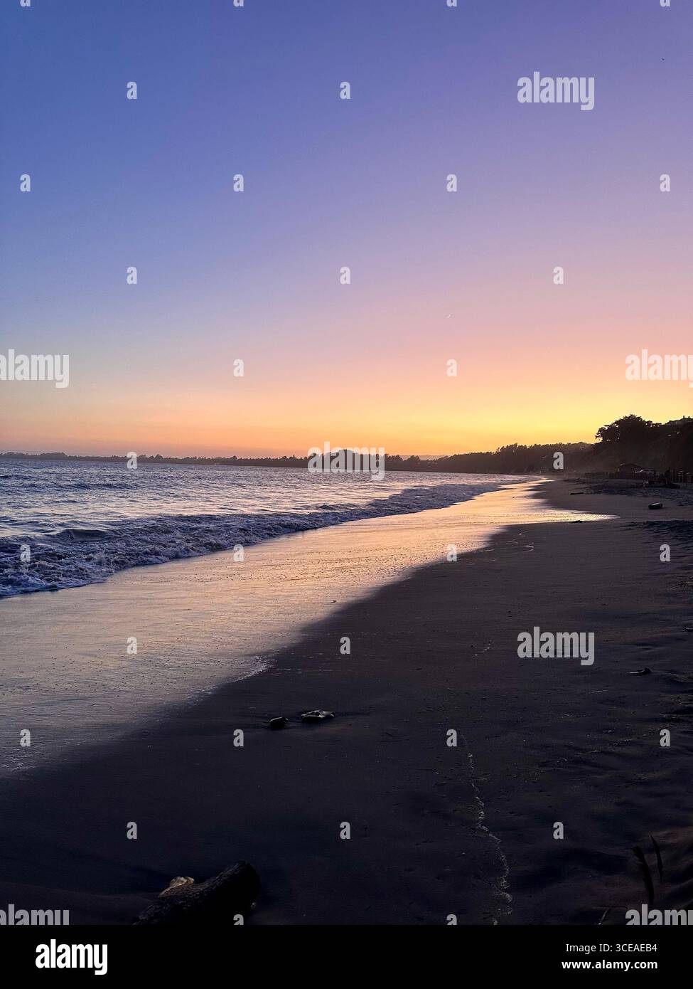 Un coucher de soleil paisible sur l'océan avec de douces vagues lavant sur la plage de sable, créant une lueur réfléchissante de teintes orange et violettes dans l'eau et le ciel. - Image de stock capturée avec un smartphone