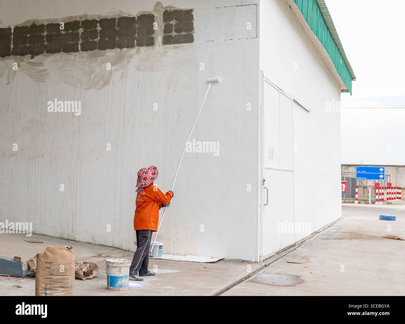 La personne porte un chapeau rouge, une chemise orange et des gants de protection. Le mur partiellement peint montre des taches de réparation de béton. Banque D'Images