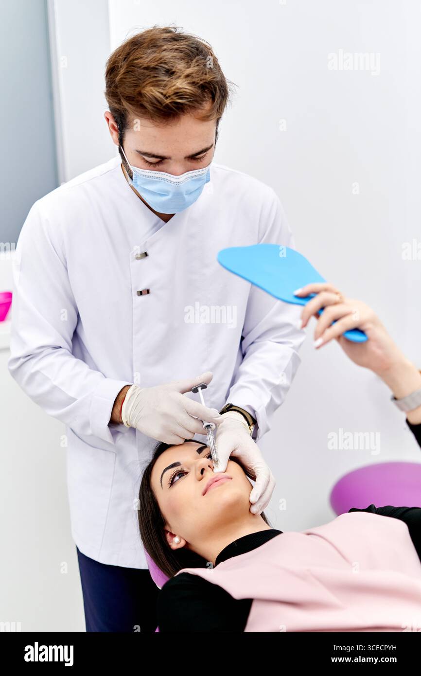 Un professionnel de la santé dans un manteau blanc et des gants stériles administre une injection d'acide hyaluronique à un patient dans un cabinet médical moderne, soulignant Banque D'Images