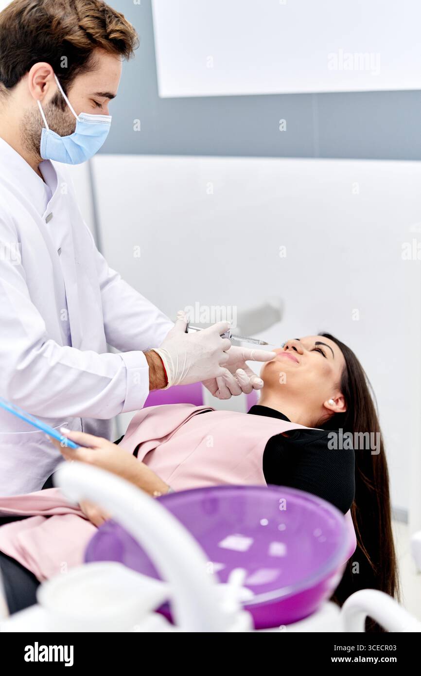 Une esthéticienne, portant un masque facial et des gants, injecte doucement de l'acide hyaluronique dans le visage d'un patient pour améliorer l'apparence de la peau dans un contexte clinique, f Banque D'Images