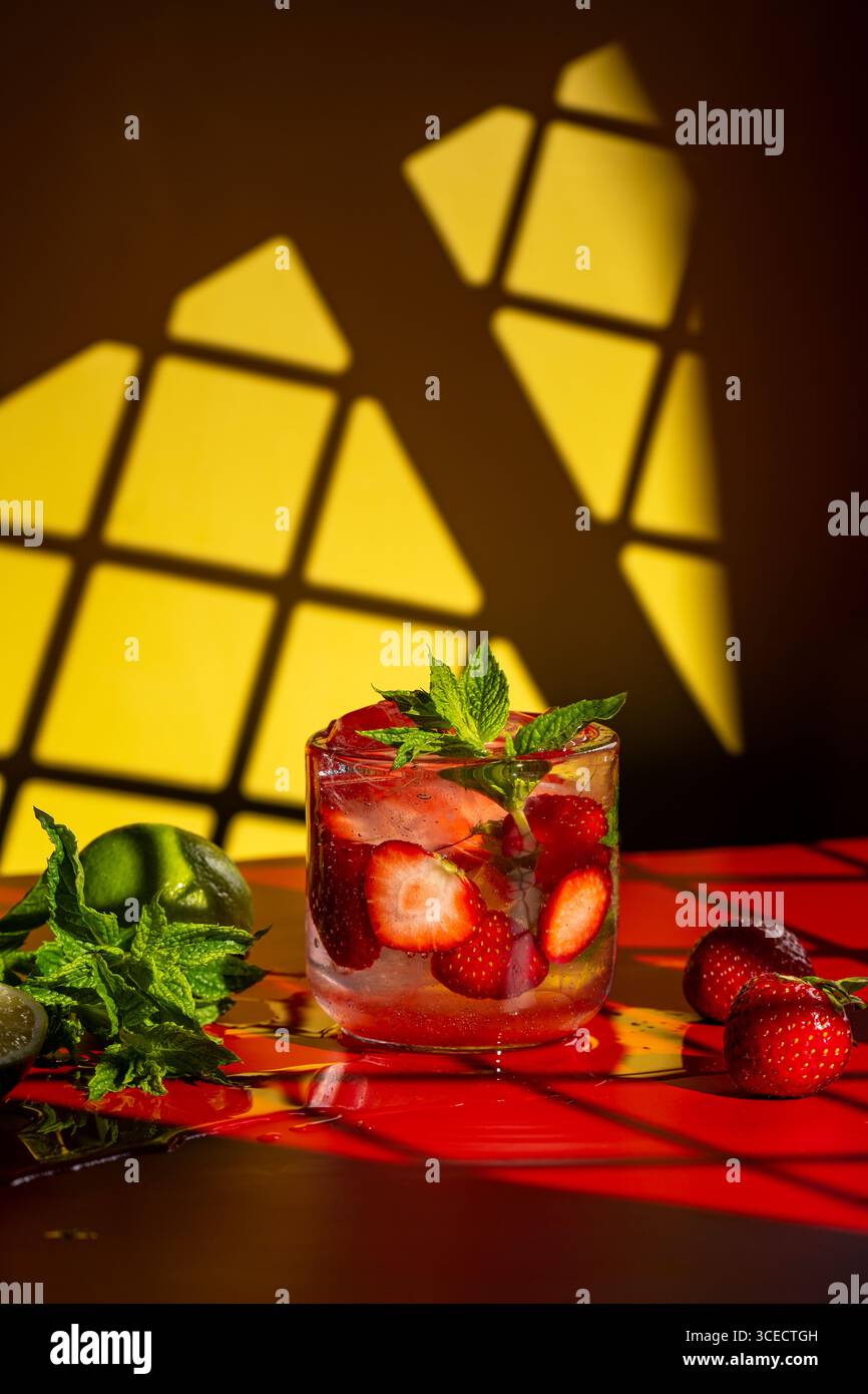 Un mojito aux fraises rafraîchissant garni de menthe fraîche et de fraises tranchées dans un verre clair. Sur une surface rouge vif, avec un citron vert et de la menthe Banque D'Images
