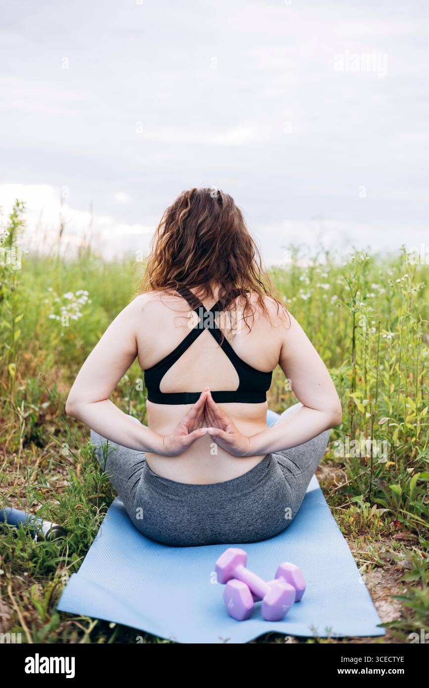 Vue arrière d'une femme méconnaissable pratiquant la pose de yoga de prière inversée dans un champ serein. Haltères violets et bouteille d'eau sur un tapis signifient fitness et Banque D'Images