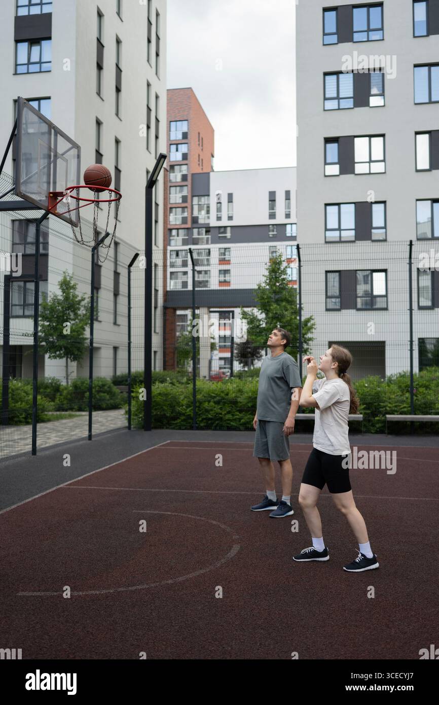 Un père et une fille jouant au basket-ball ensemble sur un terrain urbain. La fille tente de tirer pendant que le père regarde. Des bâtiments modernes entourent Banque D'Images