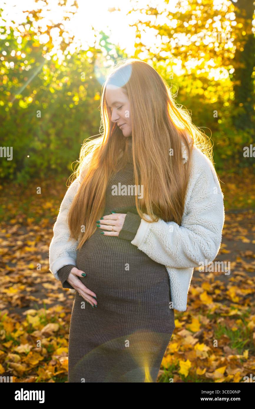 Une belle femme enceinte aux longs cheveux rouges berce son ventre, debout au milieu des feuilles dorées d'automne. La lumière du soleil filtre à travers les arbres, jetant un w Banque D'Images