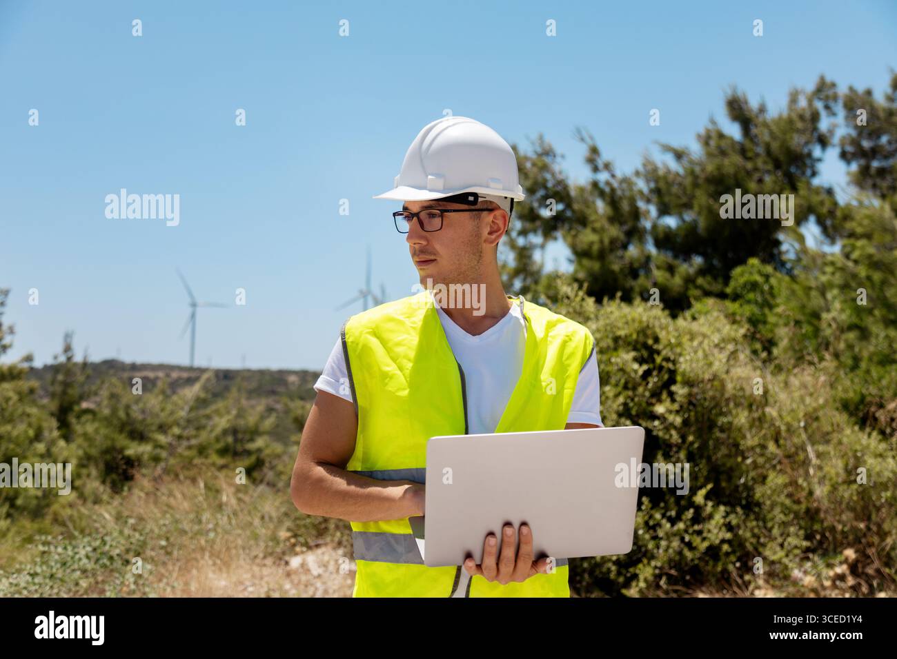 Un ingénieur dans un gilet de sécurité et un casque tient un ordinateur portable tout en inspectant les éoliennes dans une zone rurale pittoresque. Il s'assure que les turbines fonctionnent efficacement Banque D'Images