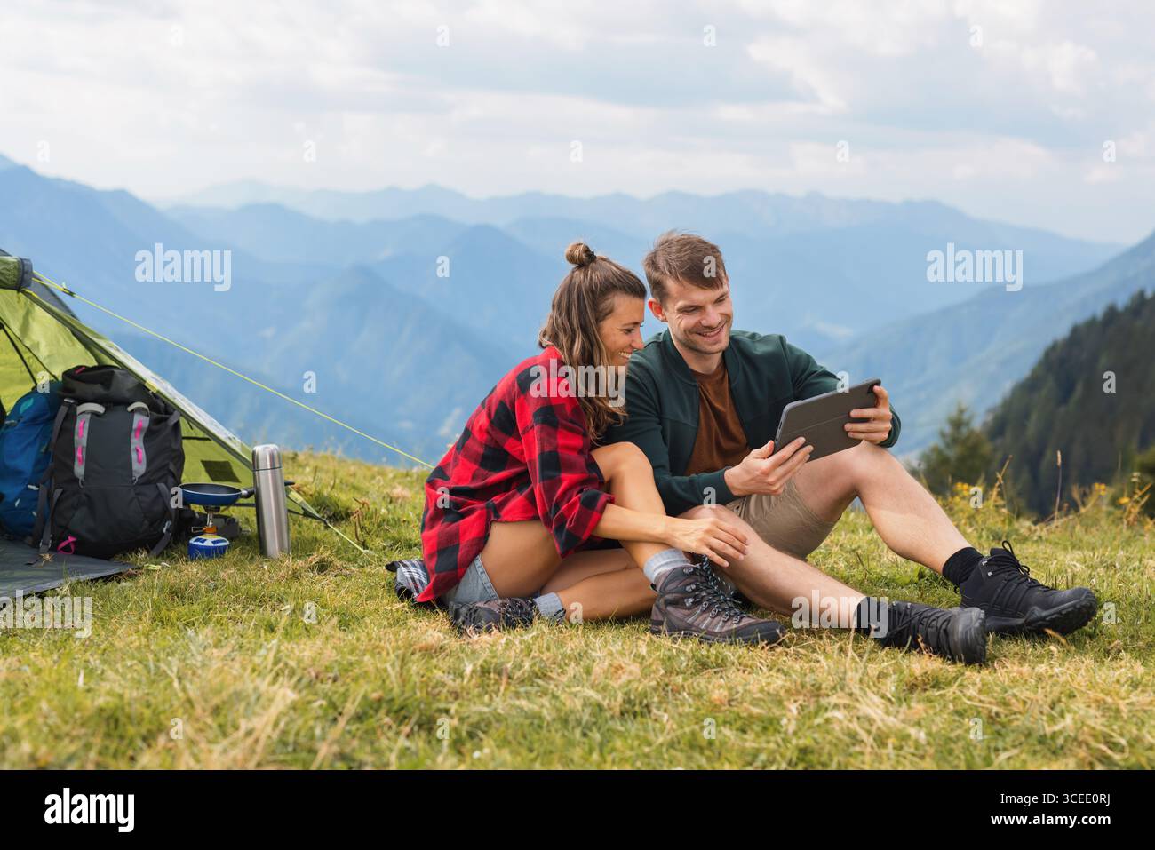Jeune couple moderne campant dans les montagnes, mélangeant technologie et nature, travaillant à distance sur une tablette tout en cuisinant à l'extérieur et en profitant de la vue. Banque D'Images