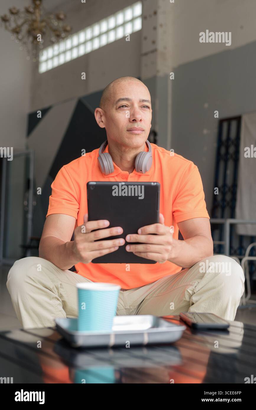 Bel homme d'âge moyen dans un polo orange à la table de café avec tasse à café, portant des écouteurs, et à l'aide d'un ordinateur tablette numérique. Technologie et cas Banque D'Images