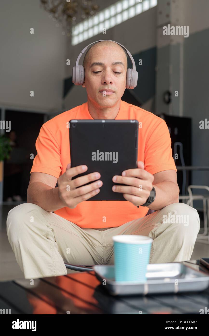 Bel homme d'âge moyen dans un polo orange à la table de café avec tasse à café, portant des écouteurs, et à l'aide d'un ordinateur tablette numérique. Technologie et cas Banque D'Images
