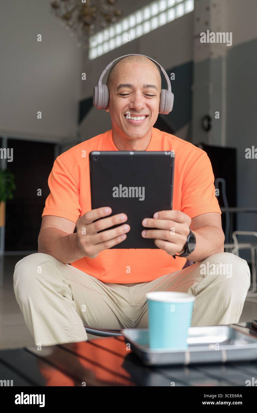 Bel homme d'âge moyen dans un polo orange à la table de café avec tasse à café, portant des écouteurs, et à l'aide d'un ordinateur tablette numérique. Technologie et cas Banque D'Images