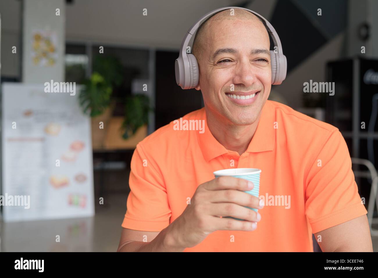 Bel homme chauve italien d'âge moyen en polo orange assis à la table du café portant des écouteurs écoutant de la musique et tenant une tasse à café. Décontracté Lif Banque D'Images