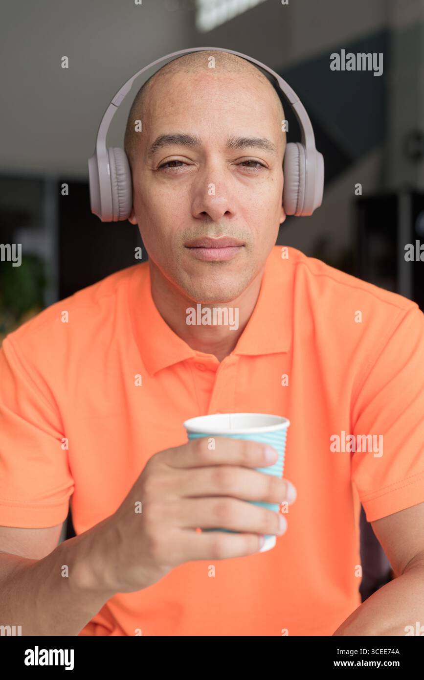 Bel homme chauve italien d'âge moyen en polo orange assis à la table du café portant des écouteurs écoutant de la musique et tenant une tasse à café. Décontracté Lif Banque D'Images