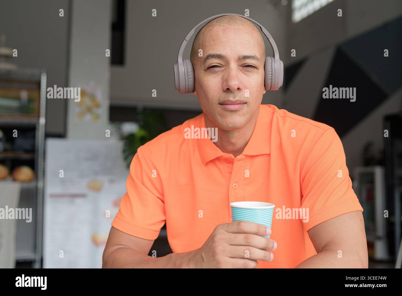Bel homme chauve italien d'âge moyen en polo orange assis à la table du café portant des écouteurs écoutant de la musique et tenant une tasse à café. Décontracté Lif Banque D'Images