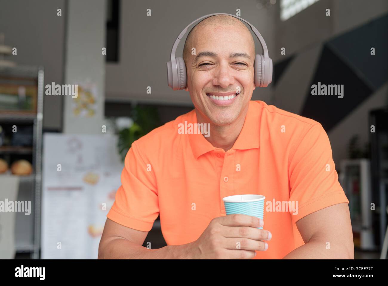 Bel homme chauve italien d'âge moyen en polo orange assis à la table du café portant des écouteurs écoutant de la musique et tenant une tasse à café. Décontracté Lif Banque D'Images