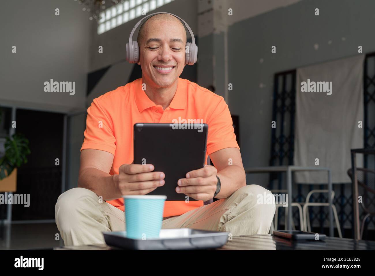 Bel homme d'âge moyen dans un polo orange à la table de café avec tasse à café, portant des écouteurs, et à l'aide d'un ordinateur tablette numérique. Technologie et cas Banque D'Images
