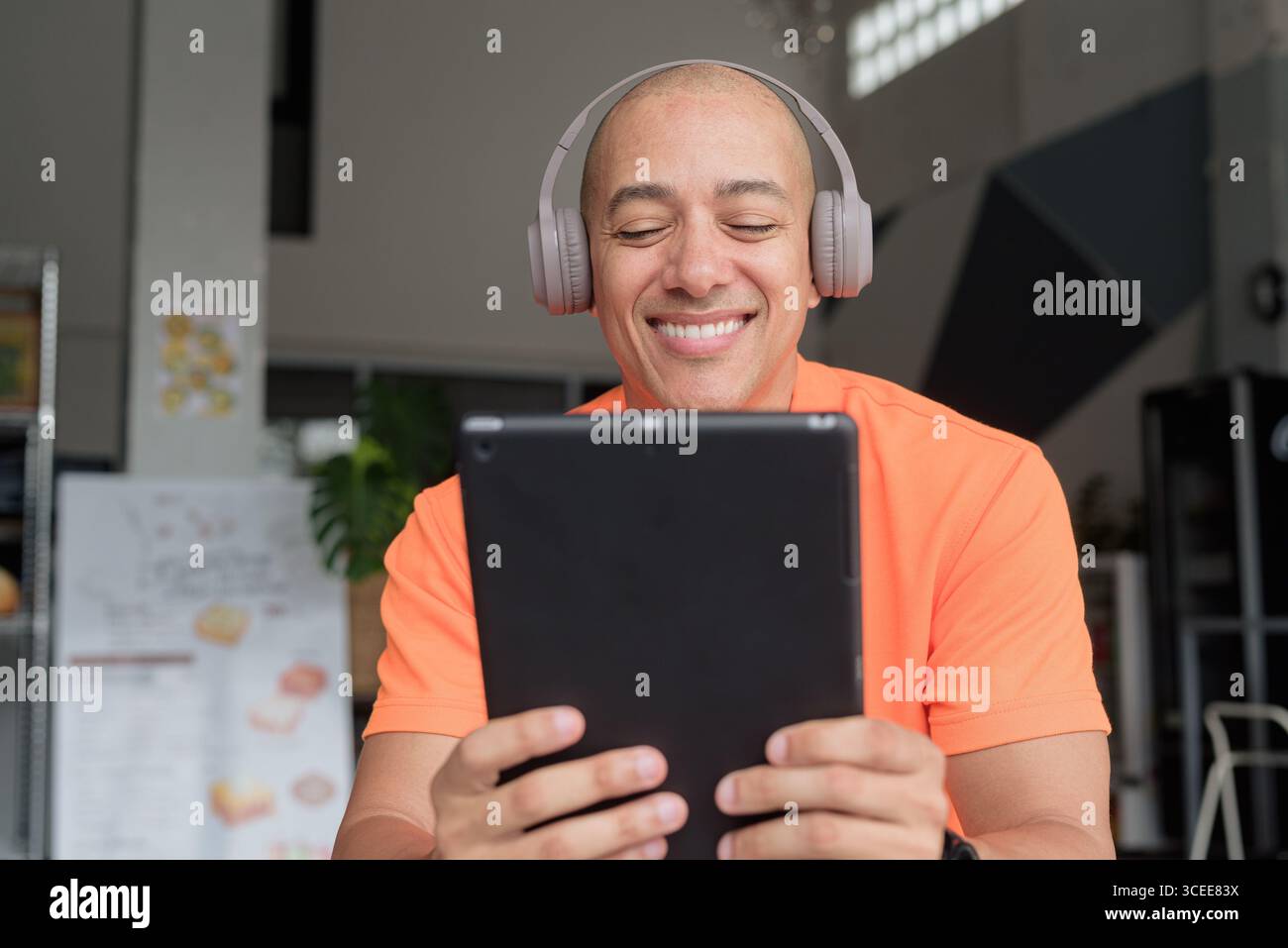 Bel homme d'âge moyen dans un polo orange à la table de café avec tasse à café, portant des écouteurs, et à l'aide d'un ordinateur tablette numérique. Technologie et cas Banque D'Images