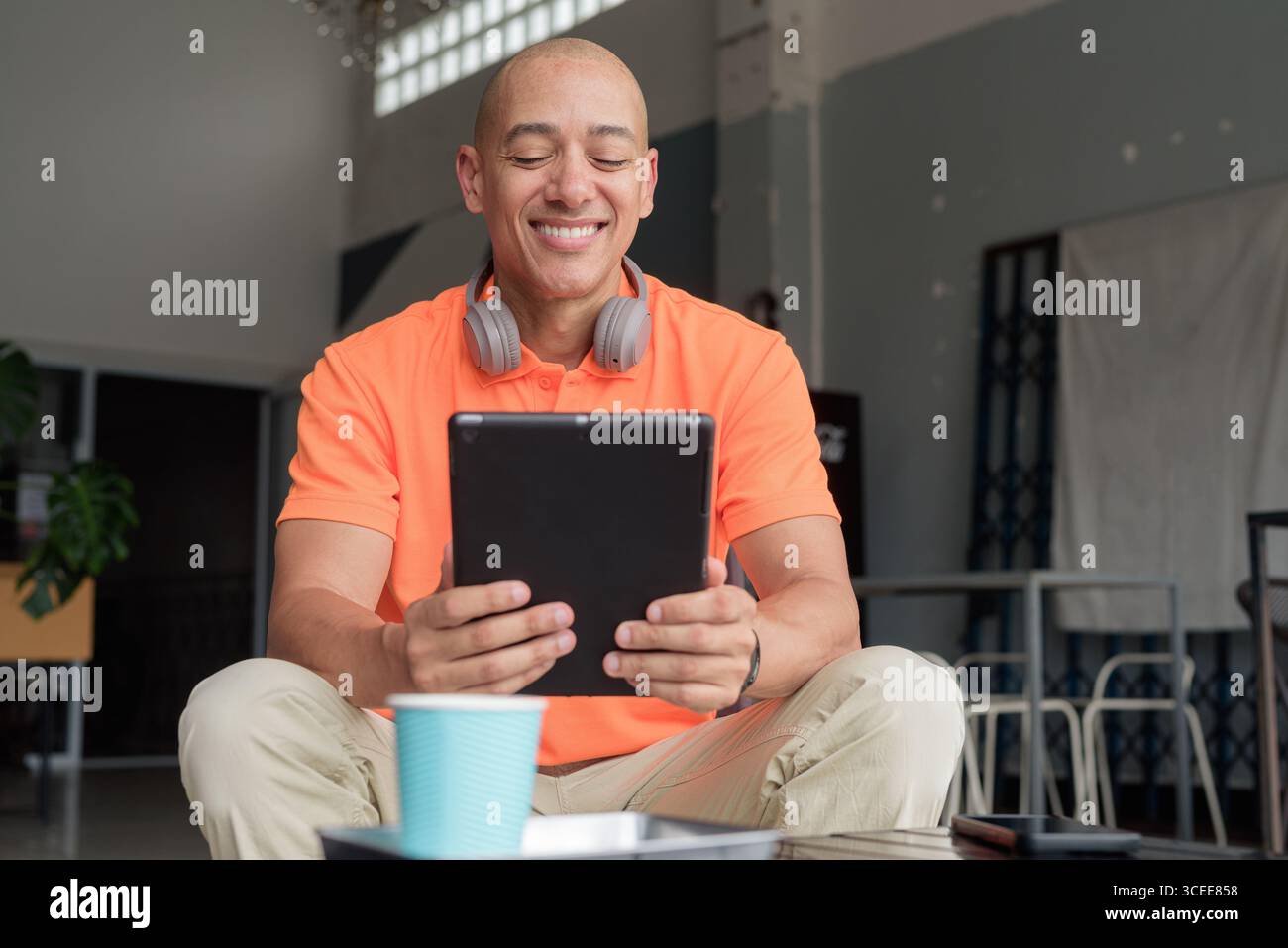Bel homme d'âge moyen dans un polo orange à la table de café avec tasse à café, portant des écouteurs, et à l'aide d'un ordinateur tablette numérique. Technologie et cas Banque D'Images