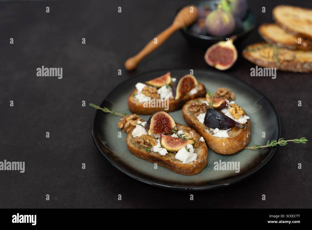 Vue de face horizontale de trois toasts de figue et fromage de chèvre sur assiette verte avec thym, noix, trempette au miel, mélange sucré salé, fond sombre Banque D'Images