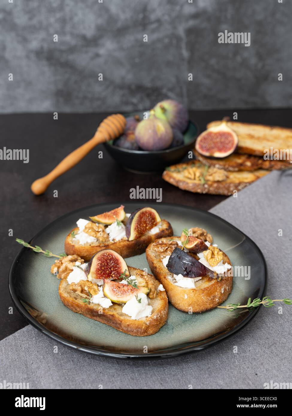 Vue de dessus verticale de trois toasts de figues et de fromage de chèvre sur une assiette verte avec un torchon gris, une trempette de miel, des figues, un espace de copie, un fond sombre Banque D'Images