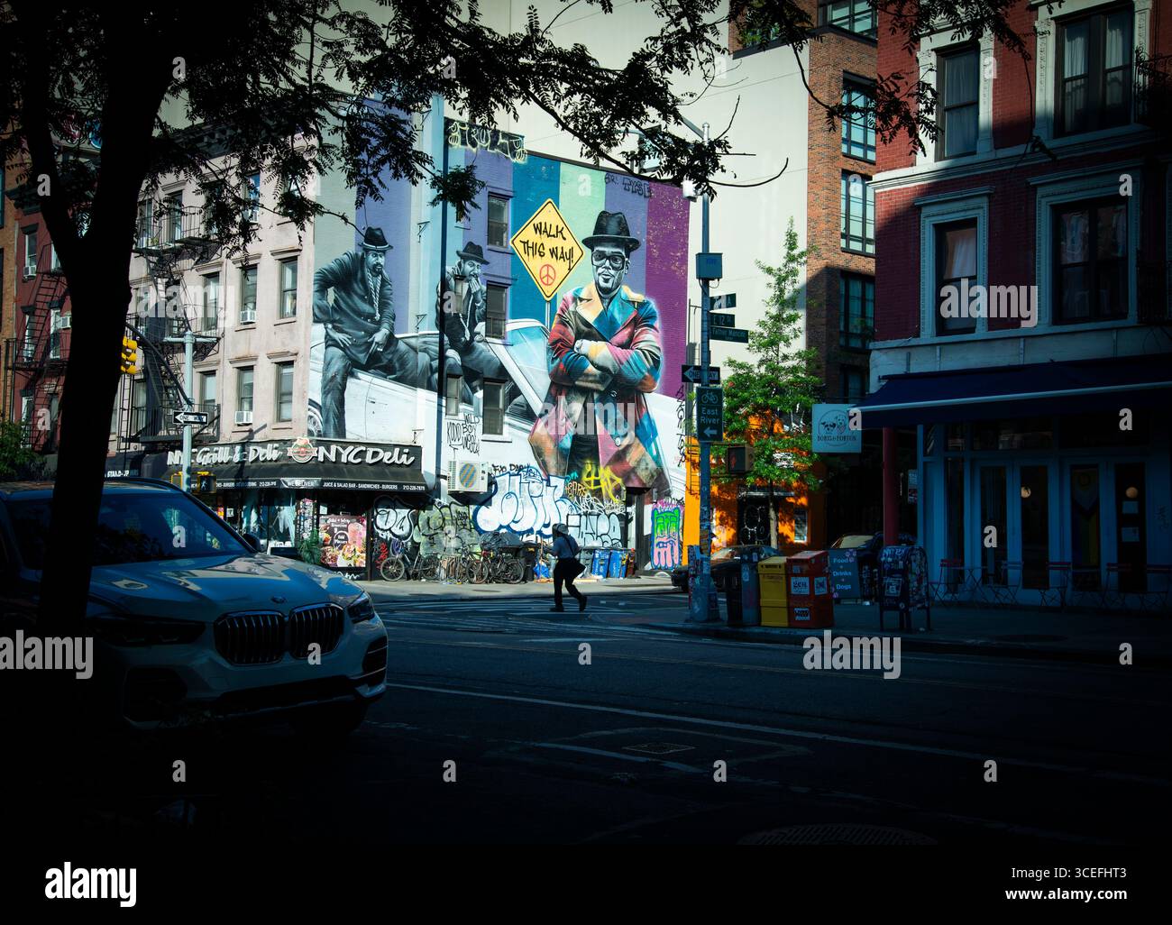 Tôt dimanche matin sur l'avenue A et East 12th Street dans l'East Village de New York. Un homme avec un café traverse East 12th Street. Une grande murale de l'artiste de rue brésilien Eduardo Kobra représentant Run-DMC, un groupe hip-hop originaire du Queens, New York, remplit le côté d'un bâtiment de quatre étages en arrière-plan. Banque D'Images