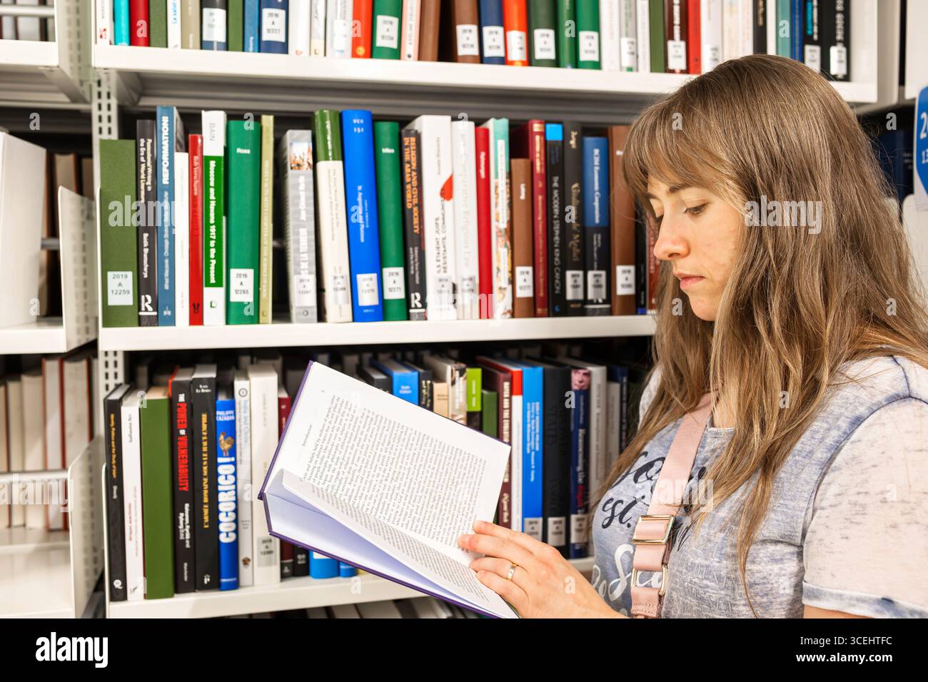 Femme caucasienne aux cheveux longs et blonds, lisant un livre à l'intérieur d'une bibliothèque publique, à côté d'étagères de livres. Banque D'Images