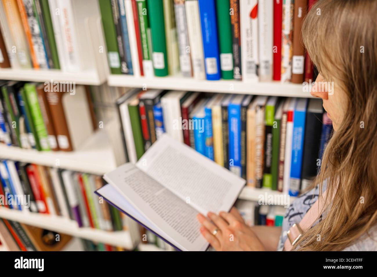 Femme caucasienne aux cheveux longs et blonds, lisant un livre à l'intérieur d'une bibliothèque publique, à côté d'étagères de livres. Banque D'Images