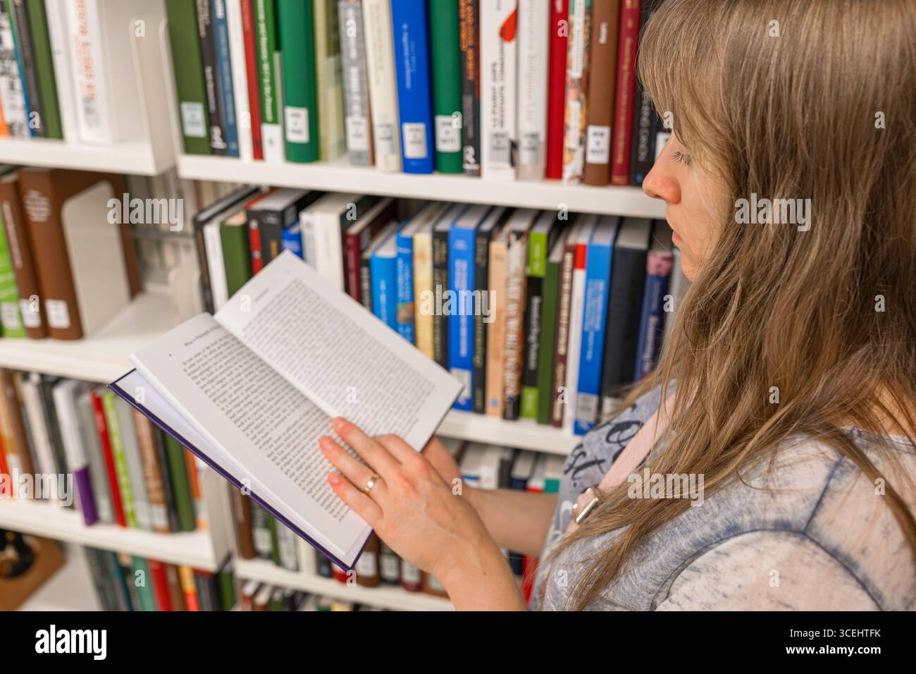 Femme caucasienne aux cheveux longs et blonds, lisant un livre à l'intérieur d'une bibliothèque publique, à côté d'étagères de livres. Banque D'Images