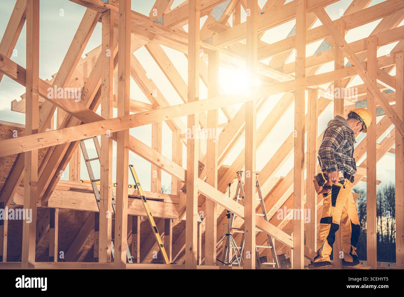 Un ouvrier de la construction assemble soigneusement un cadre en bois sous le soleil lumineux de l'après-midi. Des échelles et des outils sont dispersés autour du site actif. Banque D'Images